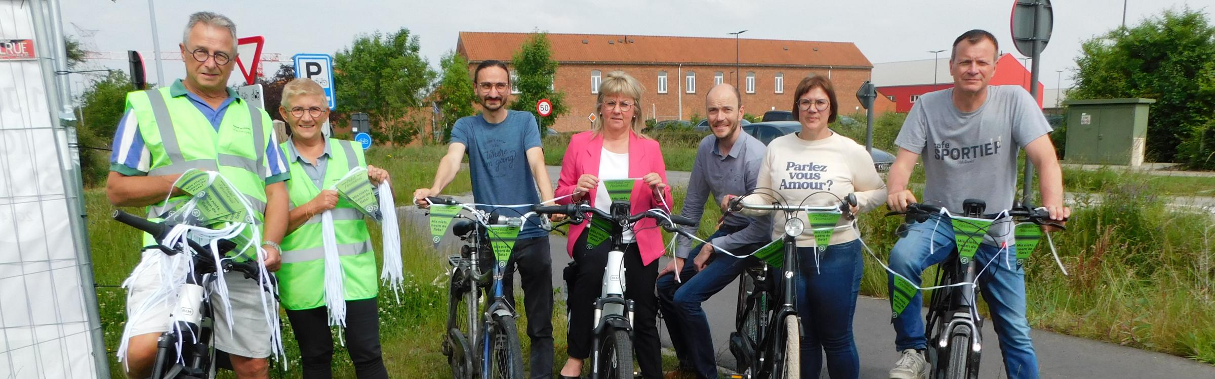 De burgemeester, schepenen en vrijwilligers delen fietsvlaggetjes uit aan passerende fietsers langs de Diksmuidse Heirweg in Zedelgem.
