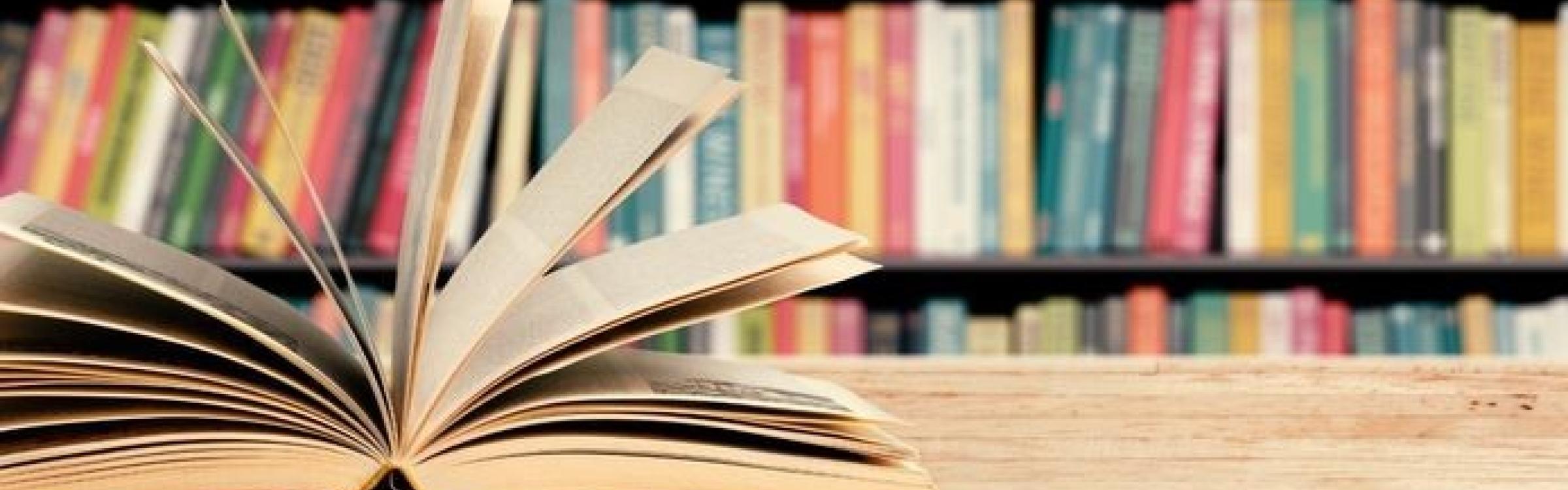 Een opengeslagen boek op een tafel en daarachter een gevuld boekenrek.