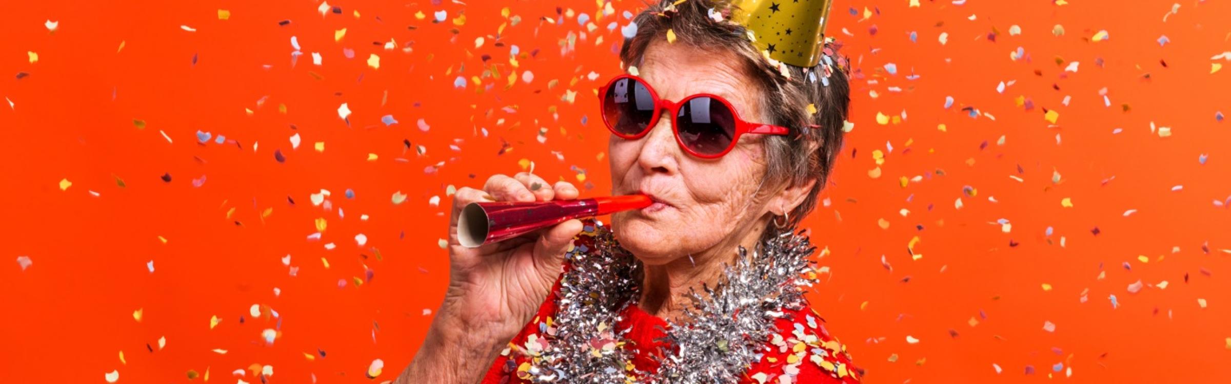 Oudere dame zit op een stoel met een feesthoedje op en blaast op een feesttoeter